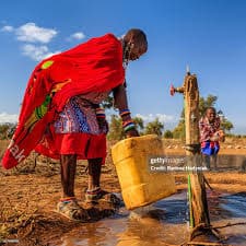 Maasai woman in red shúkà filling a jerrycan at a community tap, with a smiling child nearby—community-led water and livelihood resilience across MPIDO programmes in ASAL Kenya
